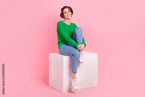 Full length photo of dreamy shiny lady wear green pullover smiling sitting white cube isolated pink color background