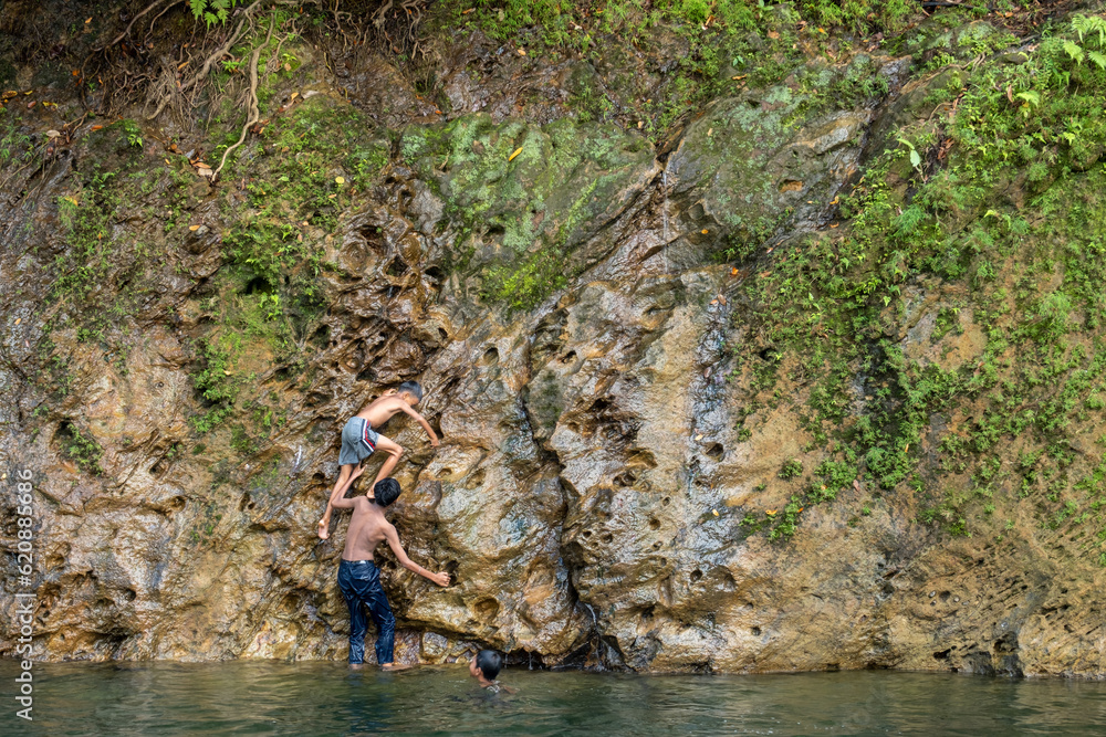 two children helping their friend to climb to the top of a cliff by the river, Indonesian boy cooperates to help his friend climb up the cliff two children helping their friend to climb to the top of a cliff by the river, Indonesian boy cooperates to help his friend climb up the cliff