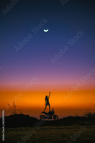 The silhouette of a girl and a motorcycle against the backdrop of a stunning sunset, and a large moon is visible in the sky.