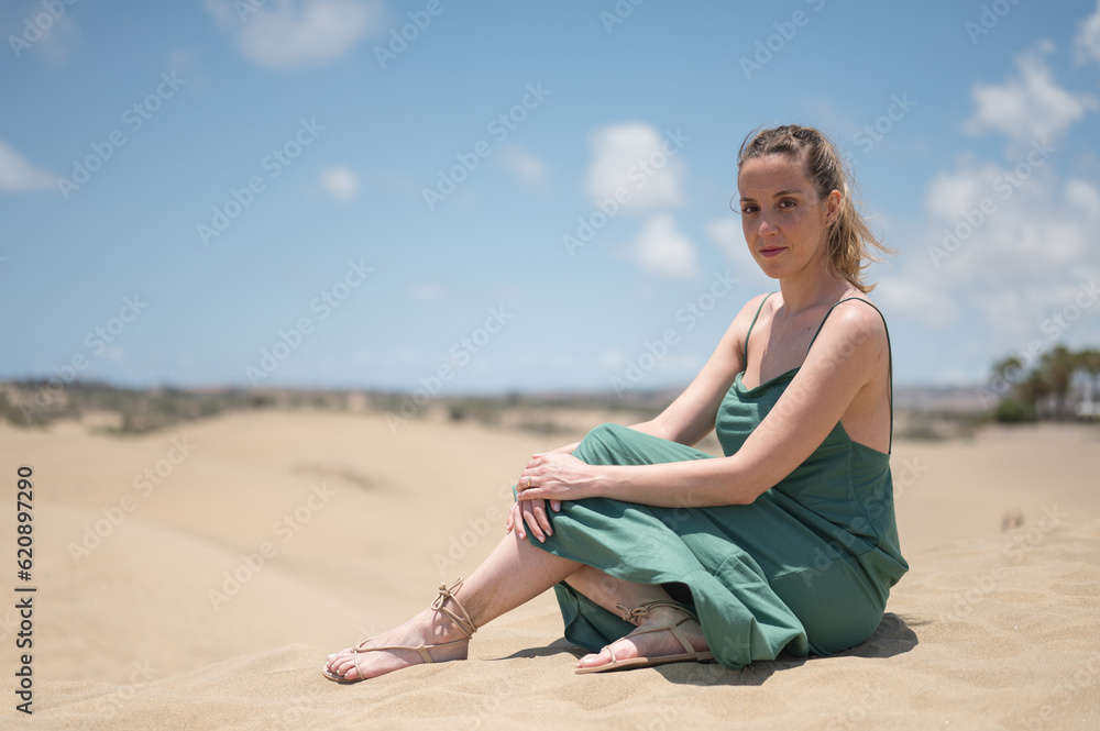 Blonde young girl in green dress sitting on sand of beach dunes