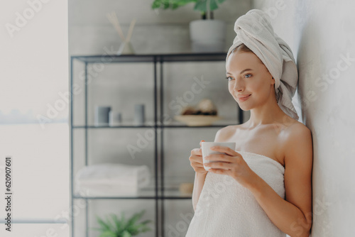 Beautiful woman with healthy skin  towel wrapped  poses with tea  concentrated  near wall in cozy home