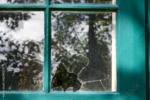 A clear transparent pane of glass in a green wood antique window broken with a hole from a gunshot. The glass has cracks, chips, and a shatter pattern. The remaining glass panes have trees reflecting. photo