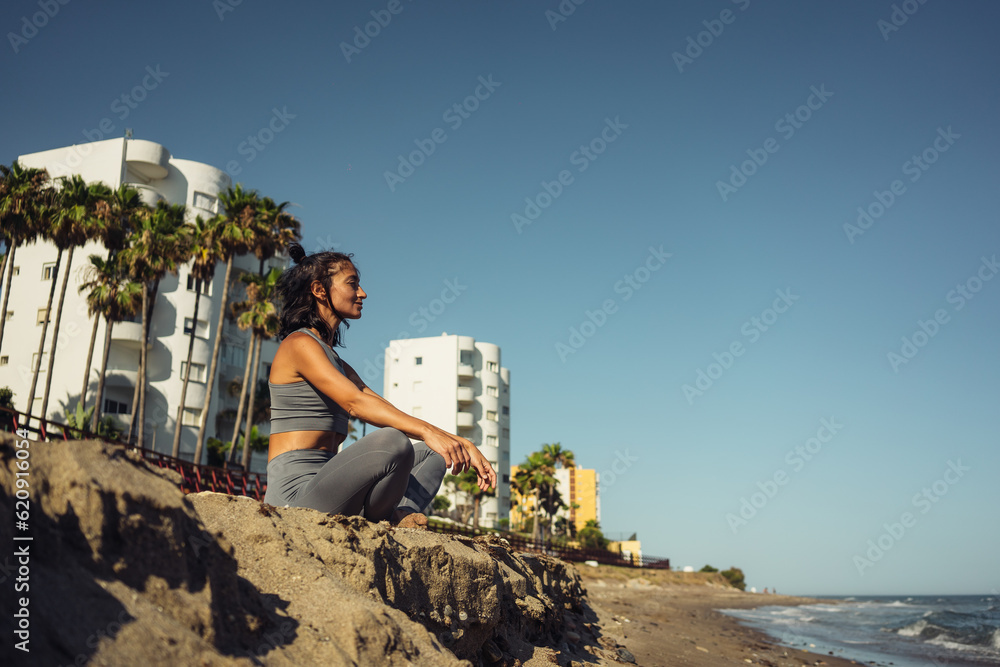 a beautiful athletic brunette girl in a gray tracksuit is engaged in meditation sitting on the sand against the backdrop of the sea