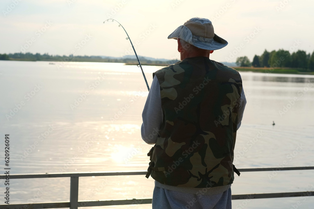 Old man fishing. Senior gray haired fisherman throws a spinning from shoreside at sunset, twisting a coil. Positive elderly male angling at lake, rotating reel rod. Retired fisher enjoying his weekend Old man fishing. Senior gray haired fisherman throws a spinning from shoreside at sunset, twisting a coil. Positive elderly male angling at lake, rotating reel rod. Retired fisher enjoying his weekend