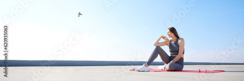 Young woman with slim, fit body sitting on fitness mantra in comfortable sportswear in sunny morning. Outdoor training