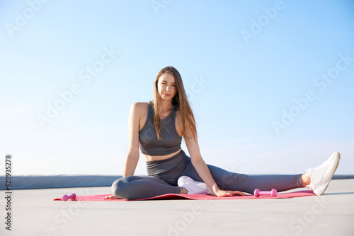 Young flexible woman, fitness trainer in comfortable sportswear training on fitness mat on warm sunny day outdoors
