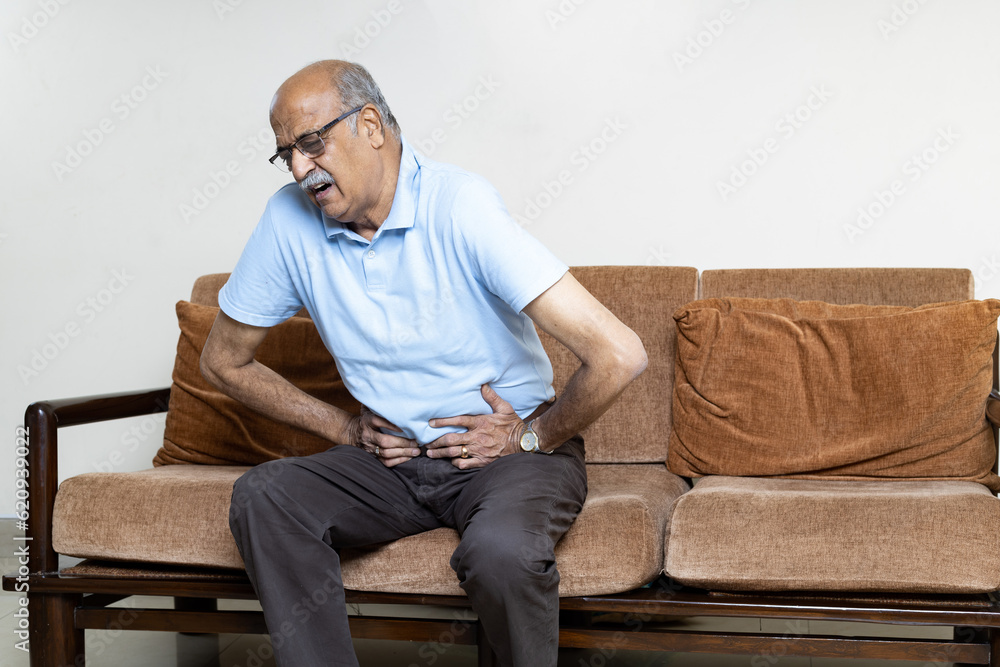 Senior men holding his stomach feeling pain while sitting on the sofa at home. Asian man having stomachache with isolated gastric. Senior suffering from digestion problem or acid reflux.