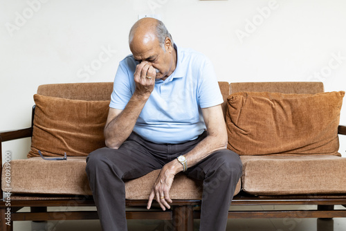 Tired, depressed senior man sitting on couch in living room feeling hurt and lonely. Aged, white-haired man touching forehead suffering from severe headache or recalling bad memories © Photographielove