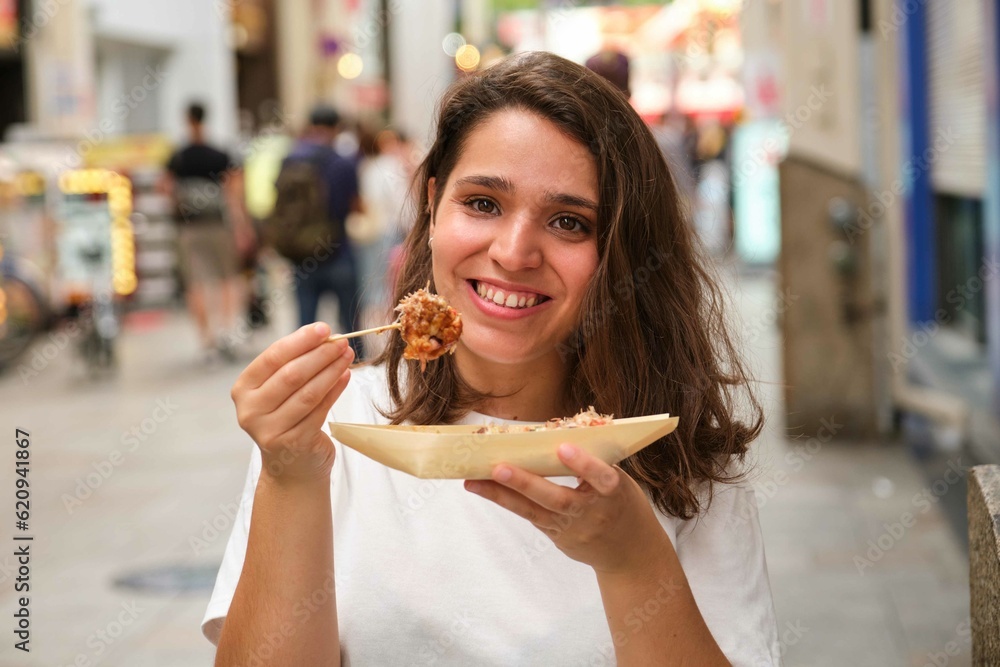 Hispanic young woman eating takoyaki and looking at camera at Dotonbori street in Osaka, Japan. Hispanic young woman eating takoyaki and looking at camera at Dotonbori street in Osaka, Japan.