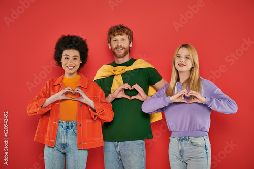 open relationship, polygamy concept, three interracial lovers showing heart sign with hands on coral background, cultural diversity, polyamorous, happy multiethnic people looking at camera photo