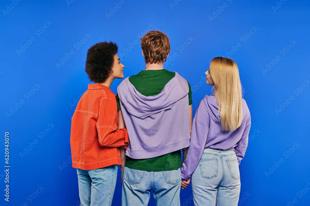polygamy, back view of polyamory three people, young man and multicultural women holding hands on blue background, studio shot, denim fashion, love triangle, bonding and love