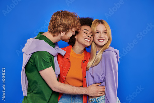 polyamory concept, positive multicultural women and redhead man hugging on blue background, studio photography, cultural diversity, polygamy, modern family, colorful clothes photo