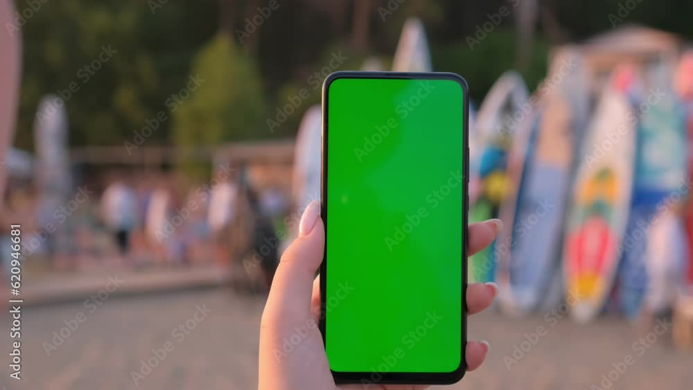 Sup rental on the beach. Female hand with smartphone with green screen mockup on tropical beach background at sunset on summer vacations Sup rental on the beach. Female hand with smartphone with green screen mockup on tropical beach background at sunset on summer vacations