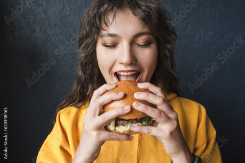 Fototapeta Attractive girl in a yellow hoodie eats a burger on a dark blue background closeup