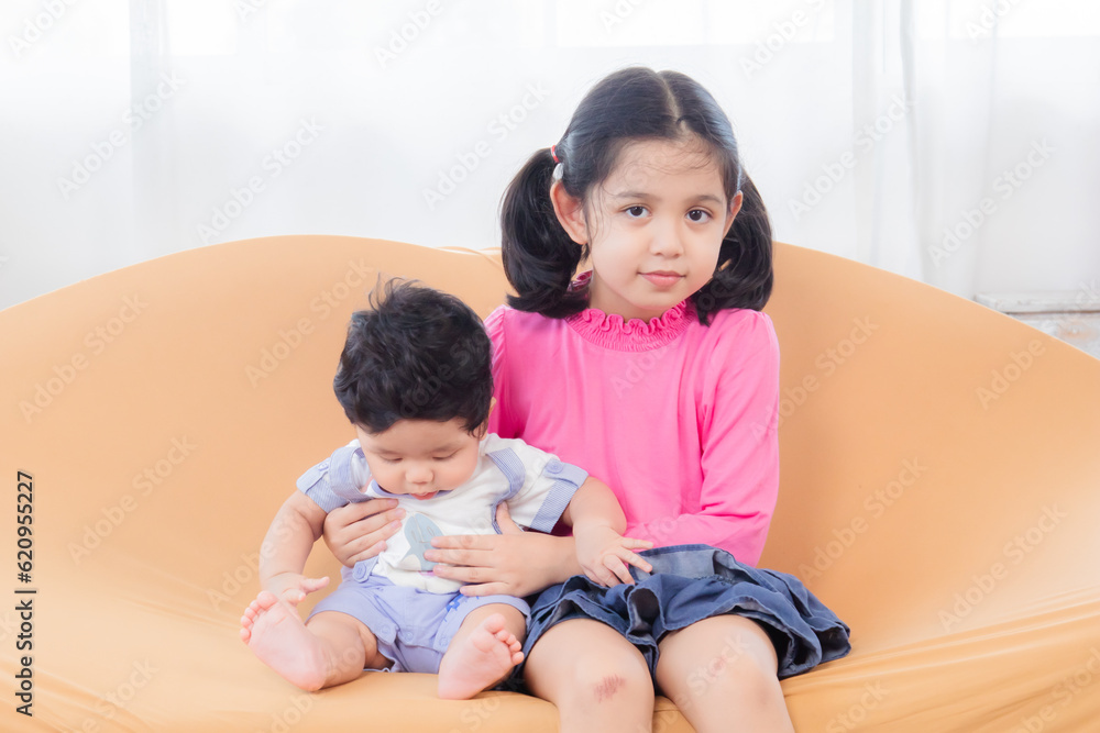Asian elder sister and newborn baby sit on sofa together, adorable infant spends time with little girl with love and care. Brothers and sisters relationship, Raising a younger brother concept. Asian elder sister and newborn baby sit on sofa together, adorable infant spends time with little girl with love and care. Brothers and sisters relationship, Raising a younger brother concept.