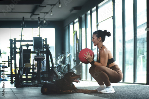 Woman exercising with exercise ball in gym and her dog lying on the floor nearby, sport woman training with fitness ball at gym
