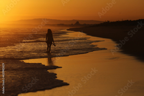 A serenidade do entardecer na praia do Algarve, em Portugal. Uma mulher caminha descalça em direção ao horizonte, aproveitando o verão a beira mar. O cenário idílico proporciona um momento de paz.