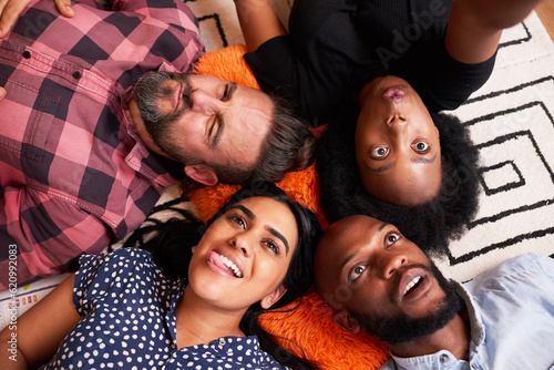 Diverse group of friends lying down taking silly selfies in circle, pulling face