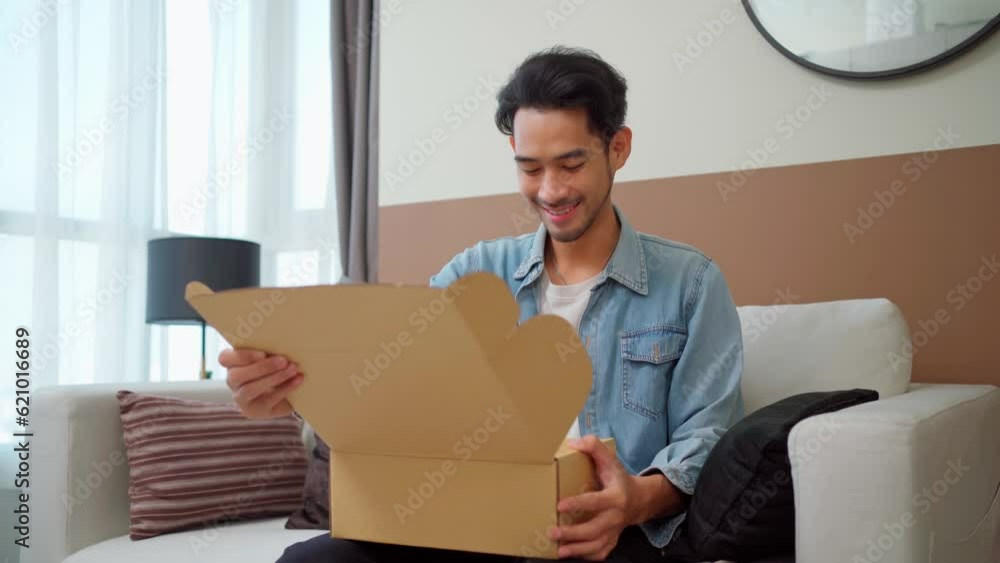 Excited young Asian man customer opening parcel box sitting at home. Smiling consumer unboxing postal shipping delivery on the sofa.