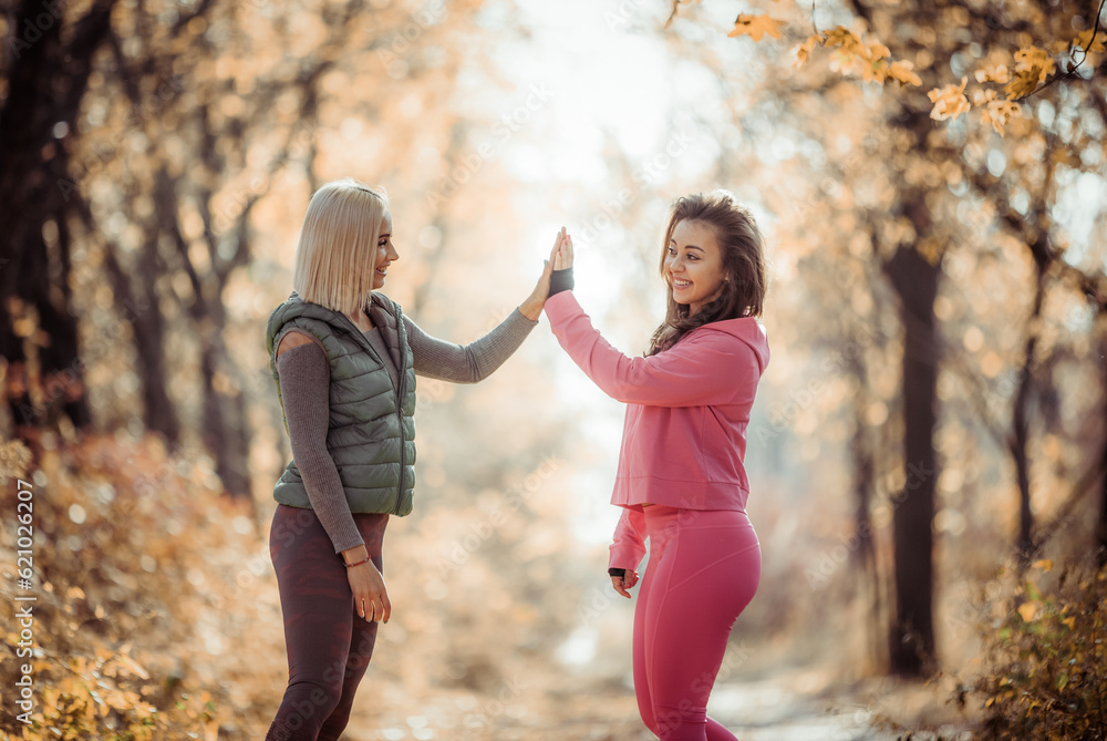 Fototapeta premium Two fitness women give five to each other in autumn forest. Healthy lifestyle