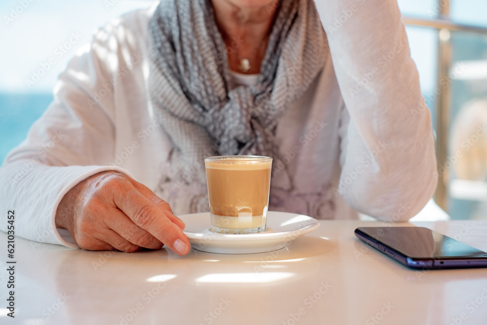 Senior woman sitting inside at coffe shop table enjoying a hot drink of coffee and milk. Smartphone on the table Senior woman sitting inside at coffe shop table enjoying a hot drink of coffee and milk. Smartphone on the table