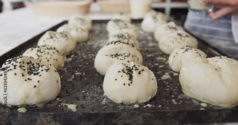 Asian baker working in bakery kitchen, sprinkling poppy seeds on rolls in slow motion