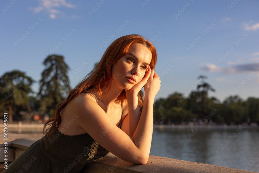 very beautiful freckled white woman with long red hair leaning on the railing and her back to the lake with the sun reflecting on her face