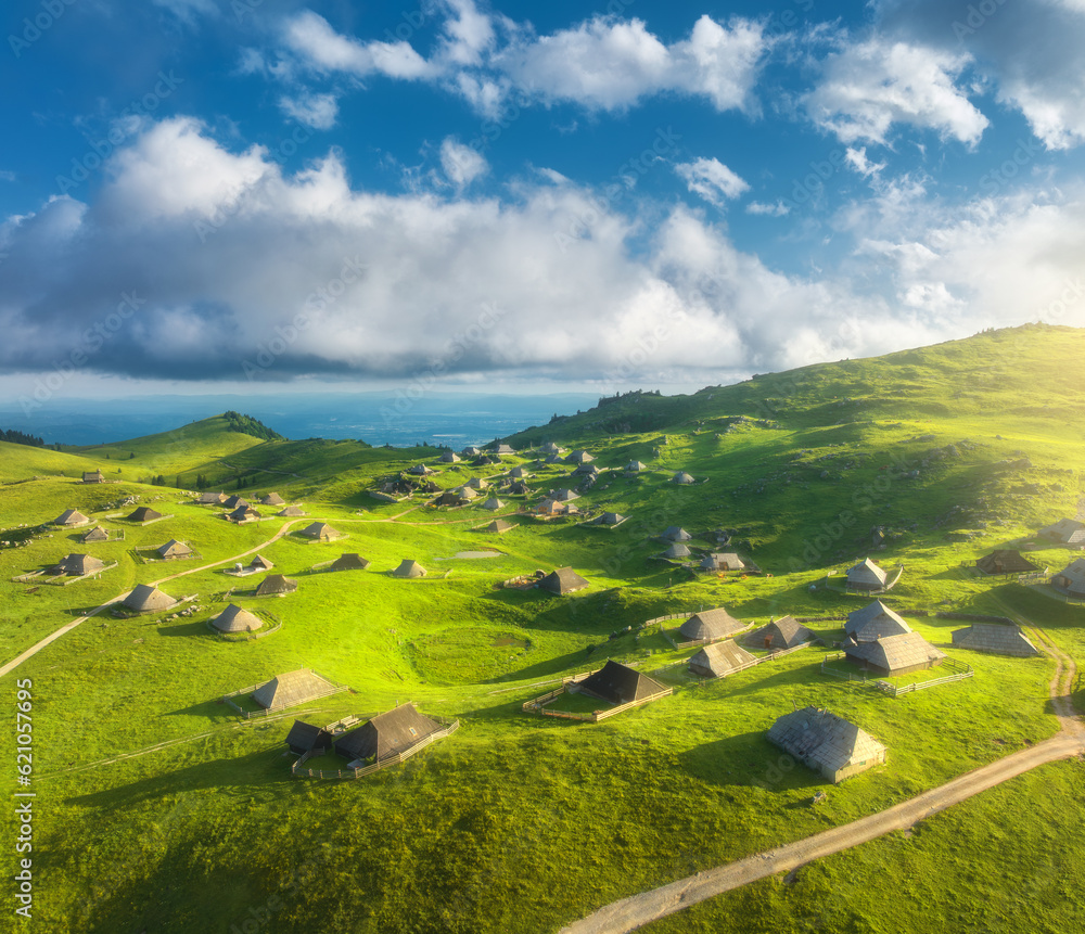 Aerial view of old mountain village and green hills at sunset in summer. Velika Planina, Slovenia. Top view of wooden houses, meadows, rural road, blue sky with clouds. Alpine shepherds settlement