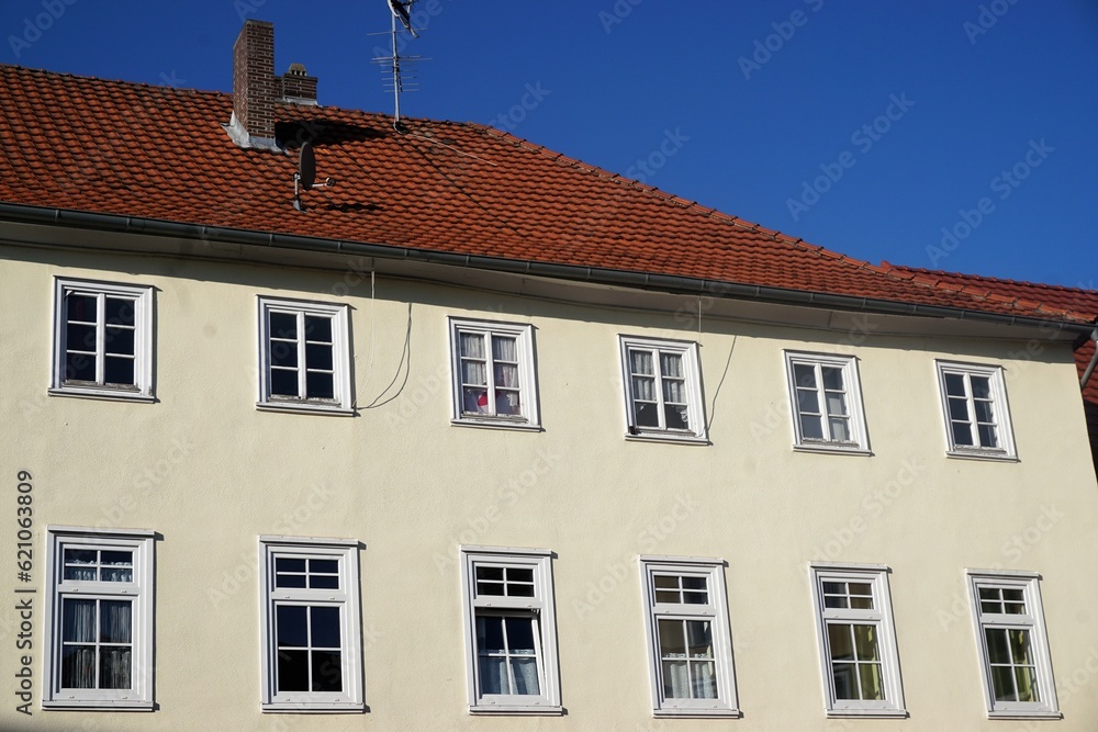 Fototapeta premium Großes altes saniertes Wohnhaus mit weißer Fassade und Fenstern mit weißen Rahmen und altem roten Ziegeldach vor blauem Himmel bei Sonne am Nachmittag im Sommer