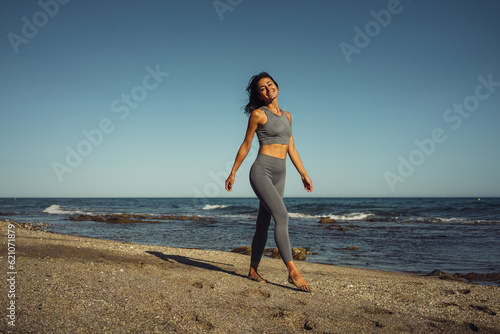 a beautiful athletic brunette girl in a gray tracksuit walks barefoot on the sand against the backdrop of the sea