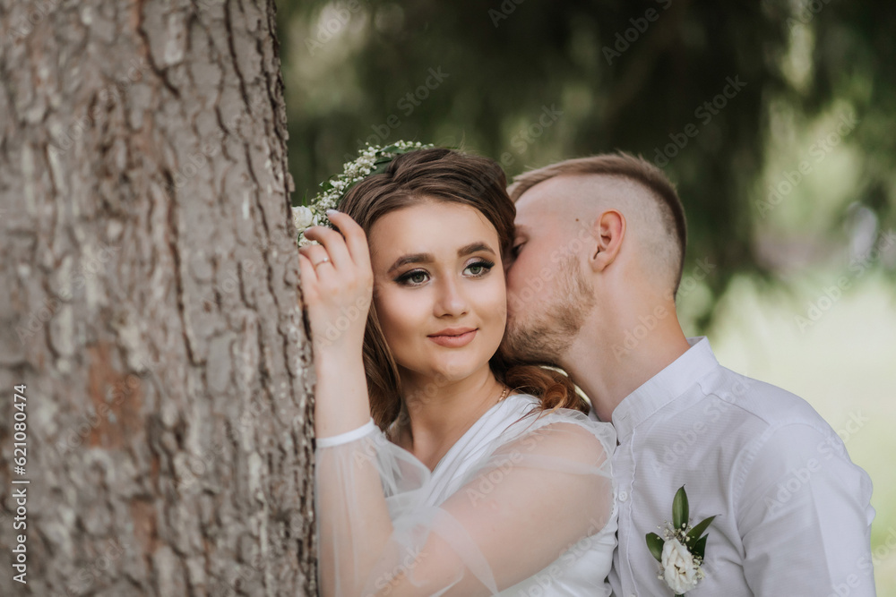 Portrait of the bride and groom. A bride with long hair and a wreath on her head near a tall tree, from behind the groom hugs her and kisses her on the cheek. Portrait of the bride and groom. A bride with long hair and a wreath on her head near a tall tree, from behind the groom hugs her and kisses her on the cheek.