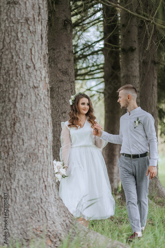the bride and groom walk in a forest with tall trees on their wedding day. The groom leads the bride by the hand in front.