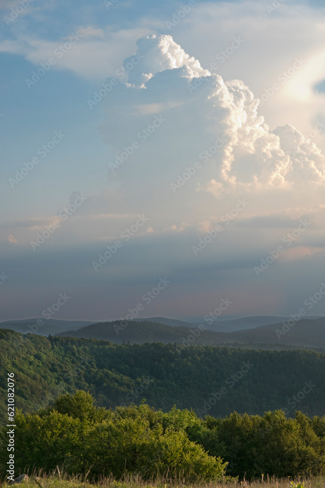 Dense, towering vertical cloud, a cumulonimbus, illuminated by the setting sun, over a wooded, hilly landscape in the Vosges