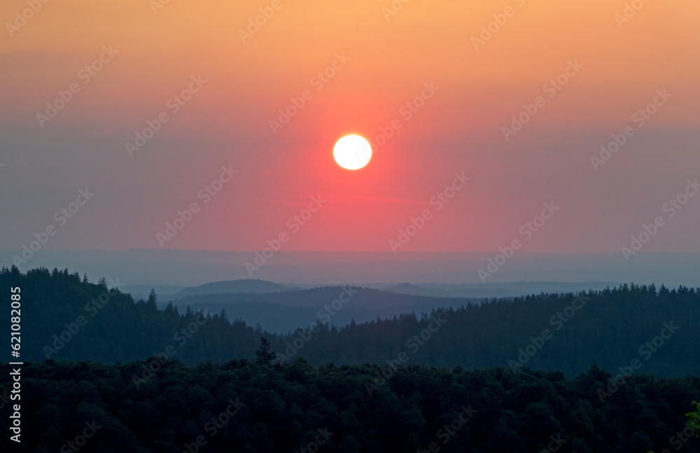 Obraz premium Sunset over de hills of the Vosges, an orange colored sun in the evening sky, hill ranges fading in the distance