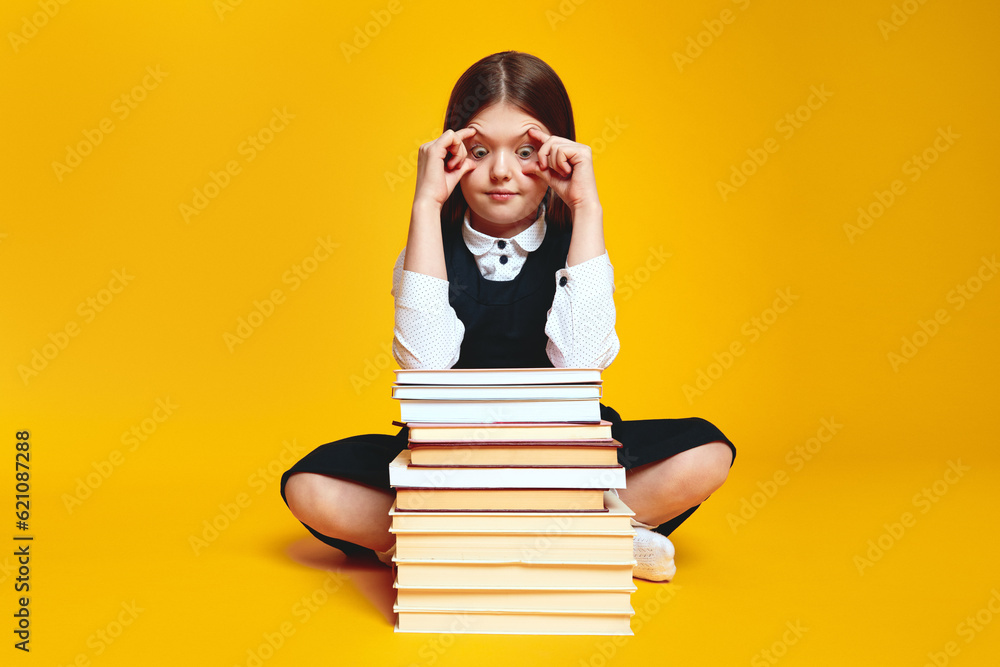 Shocked schoolgirl open eyes with fingers and looking at bunch of books, not ready for studying, sitting isolated over yellow background Shocked schoolgirl open eyes with fingers and looking at bunch of books, not ready for studying, sitting isolated over yellow background
