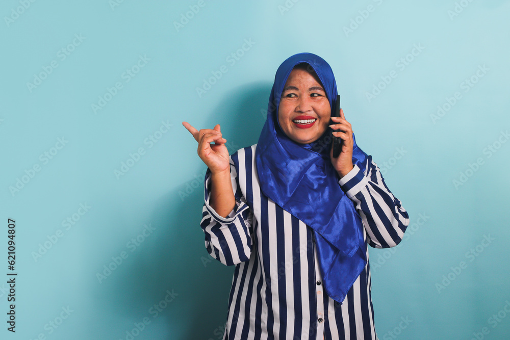 A middle-aged Asian woman in a blue hijab and a striped shirt is pointing to the left copyspace while talking on the phone. She is isolated on a blue background A middle-aged Asian woman in a blue hijab and a striped shirt is pointing to the left copyspace while talking on the phone. She is isolated on a blue background