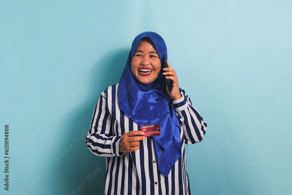 An excited middle-aged Asian woman in a blue hijab and a striped shirt is talking on the phone and showing a credit card. She is isolated on a blue background An excited middle-aged Asian woman in a blue hijab and a striped shirt is talking on the phone and showing a credit card. She is isolated on a blue background