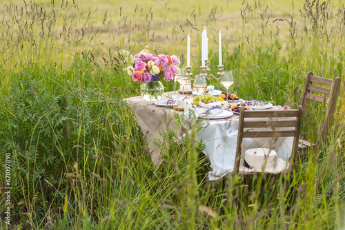 Romantic table decor for a loving couple on a meadow. Two glasses of wine, flowers in a vase, silverware, fruits, chandelier with burning candles, wooden furniture. Sunset, golden hour, summer
