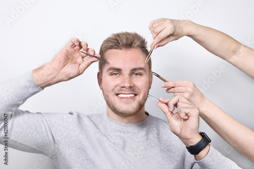 A real young blonde with blue eyes and a snow-white open smile. Two pairs of hands guide an eyebrow tweezer to his face as he trims his eyebrows. Cosmetology for men, close-up
