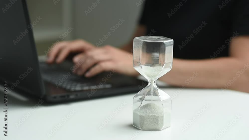 Businesswoman is working typing on a laptop, hands and a PC view are sitting at the workplace, in the foreground is a close-up of an hourglass showing the passage of time. Businesswoman is working typing on a laptop, hands and a PC view are sitting at the workplace, in the foreground is a close-up of an hourglass showing the passage of time.
