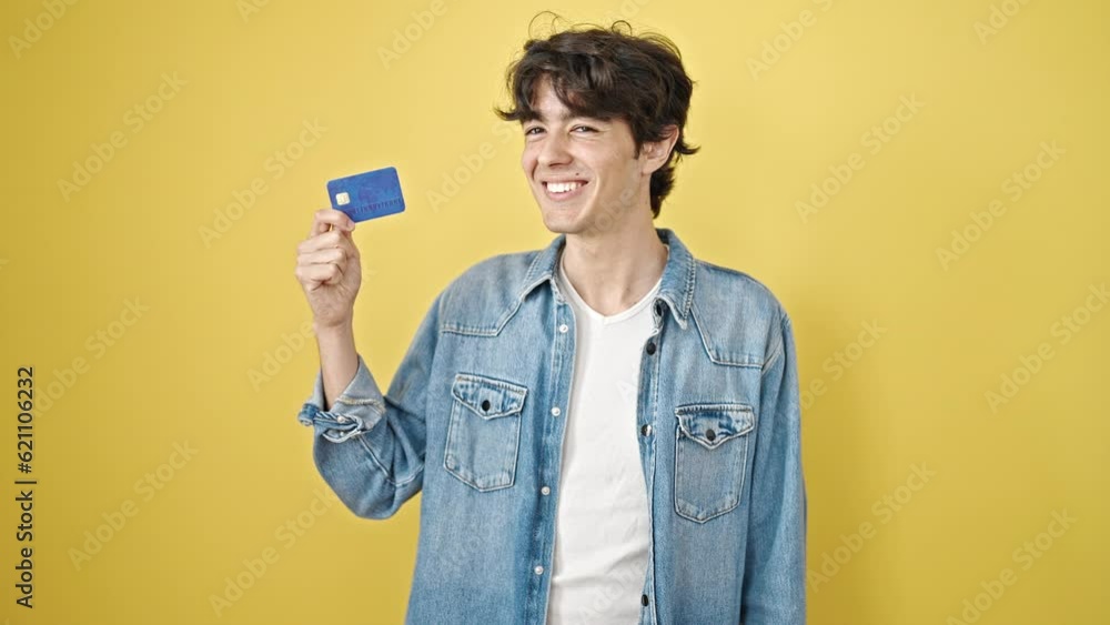 Young hispanic man holding credit card with winner expression over isolated yellow background Young hispanic man holding credit card with winner expression over isolated yellow background