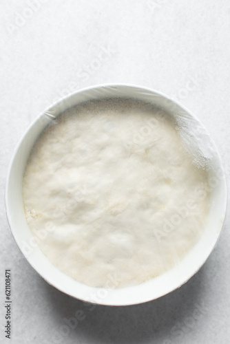 Plastic wrap covered bread dough that has doubled in size , dough that has fermented, proofed dough in a white bowl , process of making focaccia