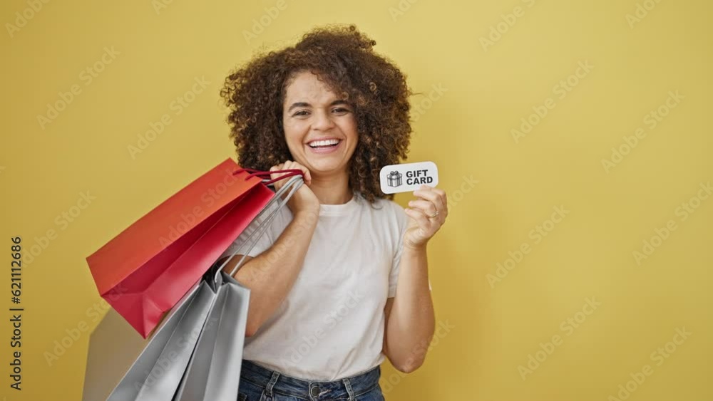 Young beautiful hispanic woman holding shopping bags and gift card smiling over isolated yellow background Young beautiful hispanic woman holding shopping bags and gift card smiling over isolated yellow background