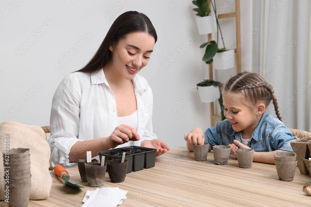 Fototapeta premium Mother and her daughter planting vegetable seeds into peat pots with soil at wooden table indoors