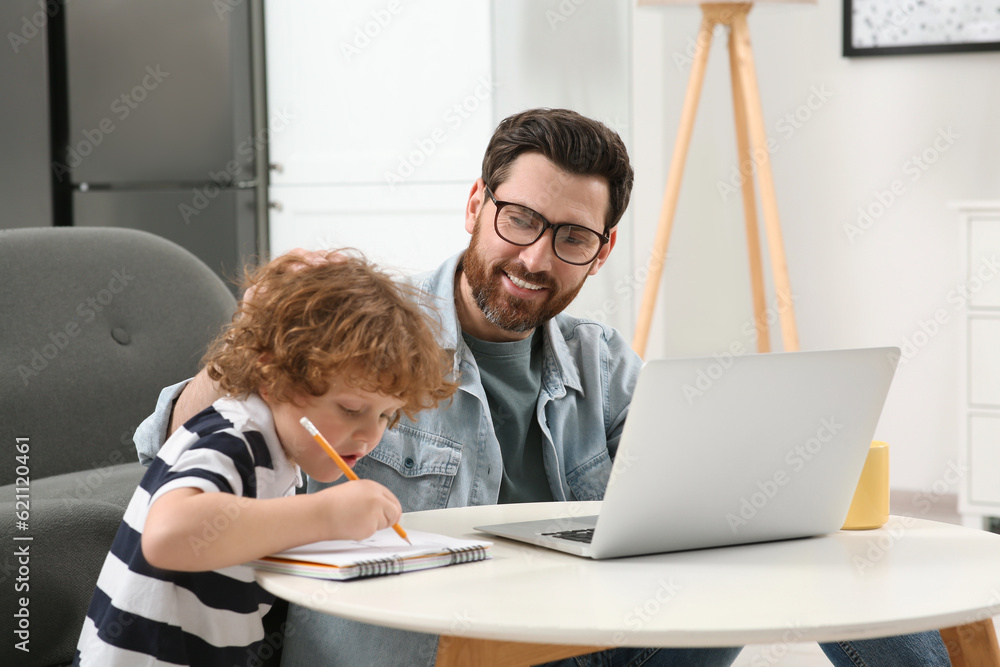 Man working remotely at home. Father using laptop while his son drawing at desk Man working remotely at home. Father using laptop while his son drawing at desk