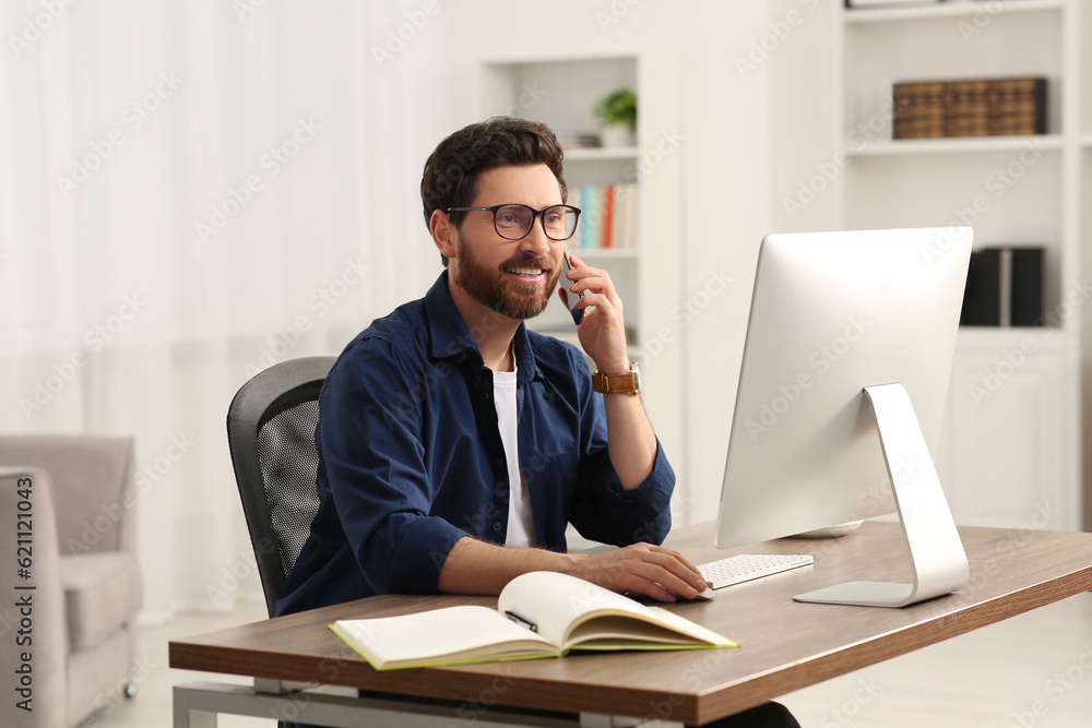 Home workplace. Happy man talking on smartphone while working with computer at wooden desk in room Home workplace. Happy man talking on smartphone while working with computer at wooden desk in room