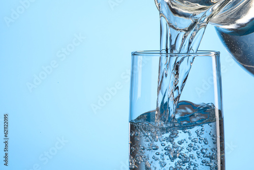Pouring water from jug into glass on light blue background, closeup. Space for text