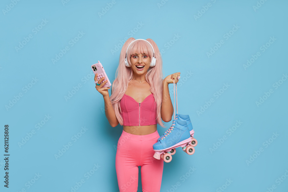 Barbie style woman in pink top and leggins with blue roller-skates, phone and white headphones posing on blue background, vintage fashion concept, copy space Barbie style woman in pink top and leggins with blue roller-skates, phone and white headphones posing on blue background, vintage fashion concept, copy space