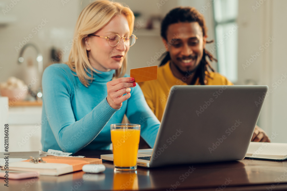 A cheerful interracial couple is sitting at their cozy home and using a credit card for online bill payment on the laptop. A cheerful interracial couple is sitting at their cozy home and using a credit card for online bill payment on the laptop.