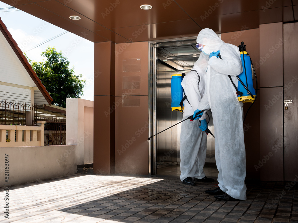 Asian teamwork man use sanitizer gun spray on the floor outdoor while wearing safety uniform, hygiene face mask, goggle and chemical backpack at the office company with elevator background